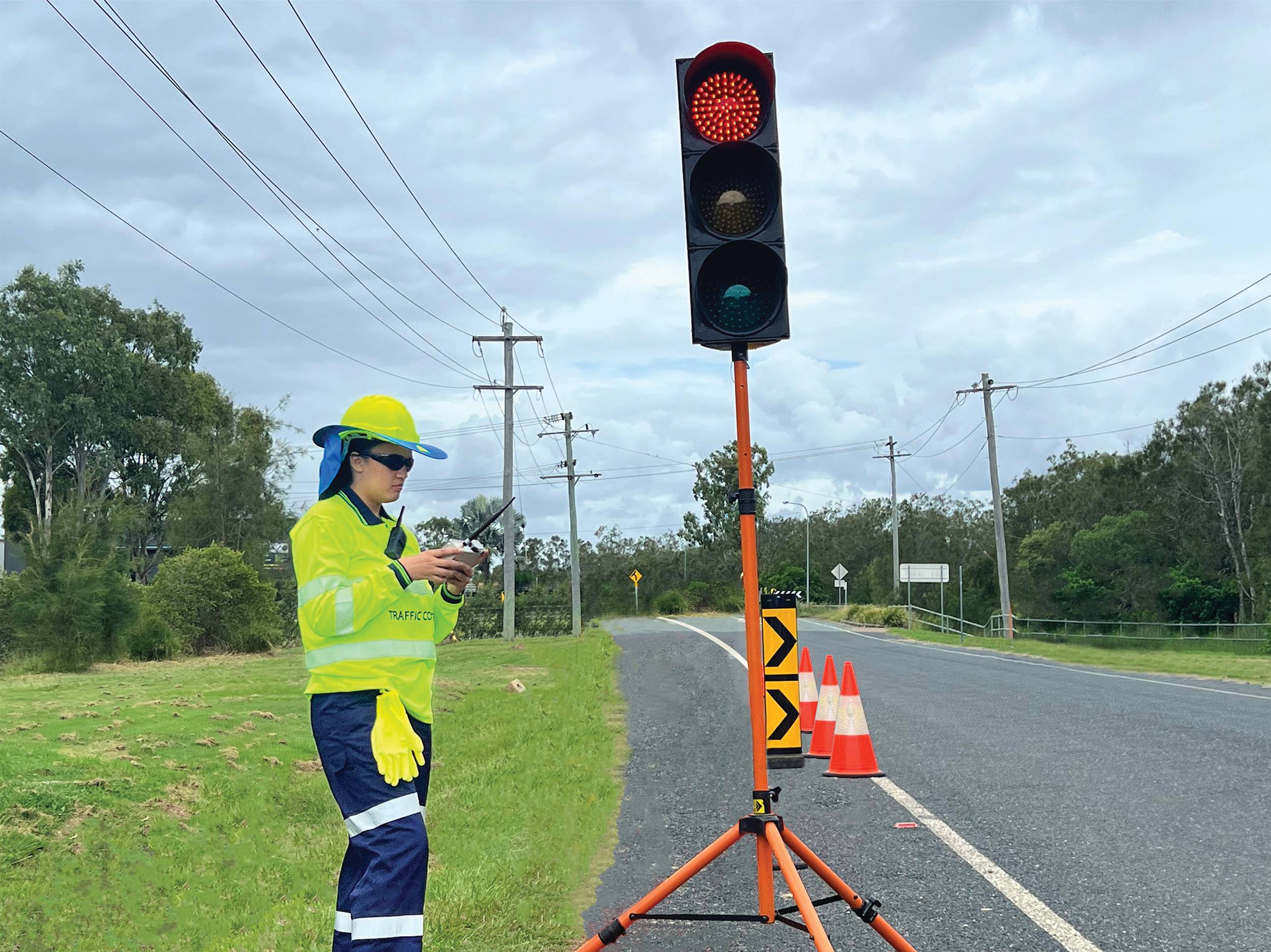 A traffic controller in high visibility clothing uses a remote to operate a portable traffic light on a rural road.