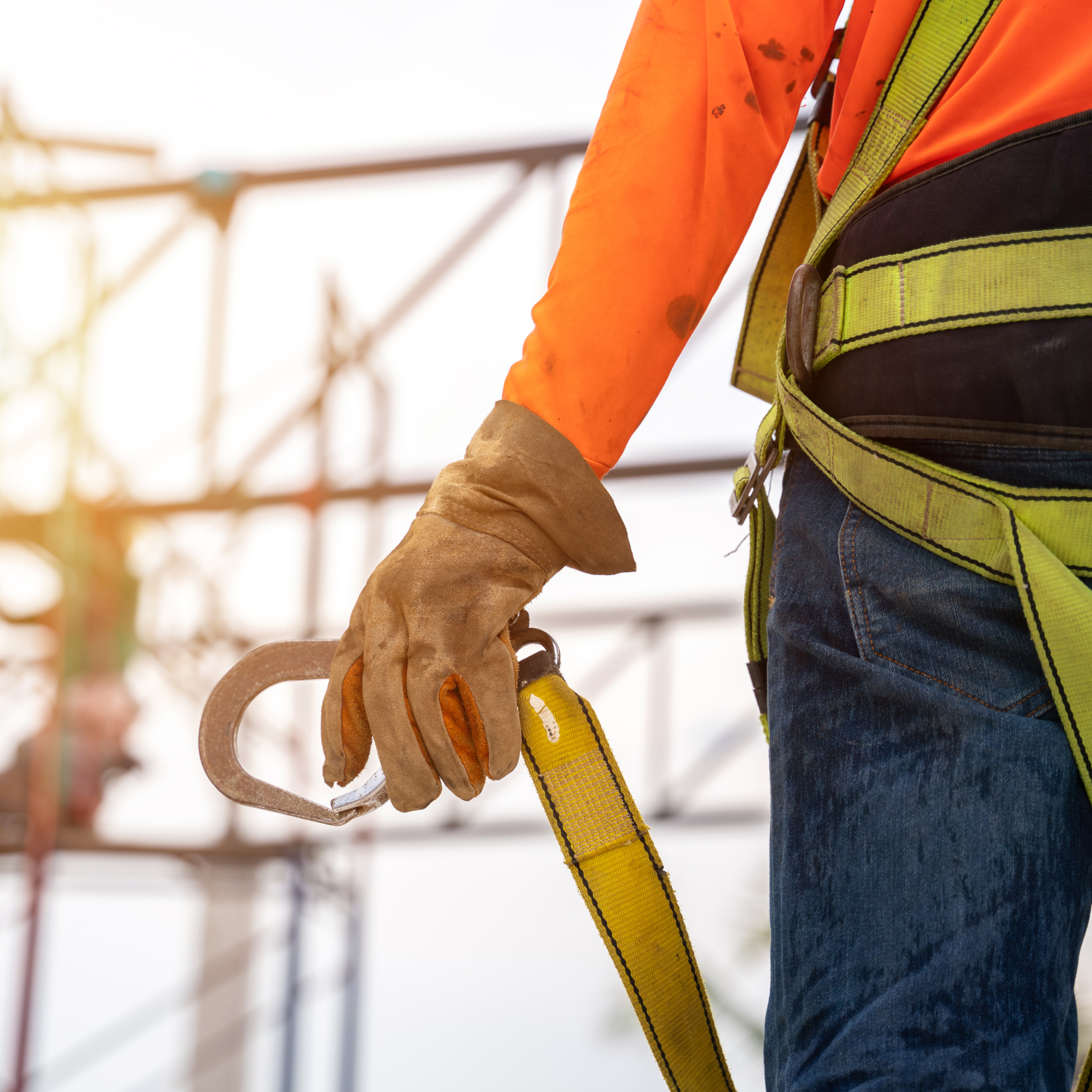 Construction worker in safety gear holding a carabiner attached to a yellow safety harness, with scaffolding in the background.