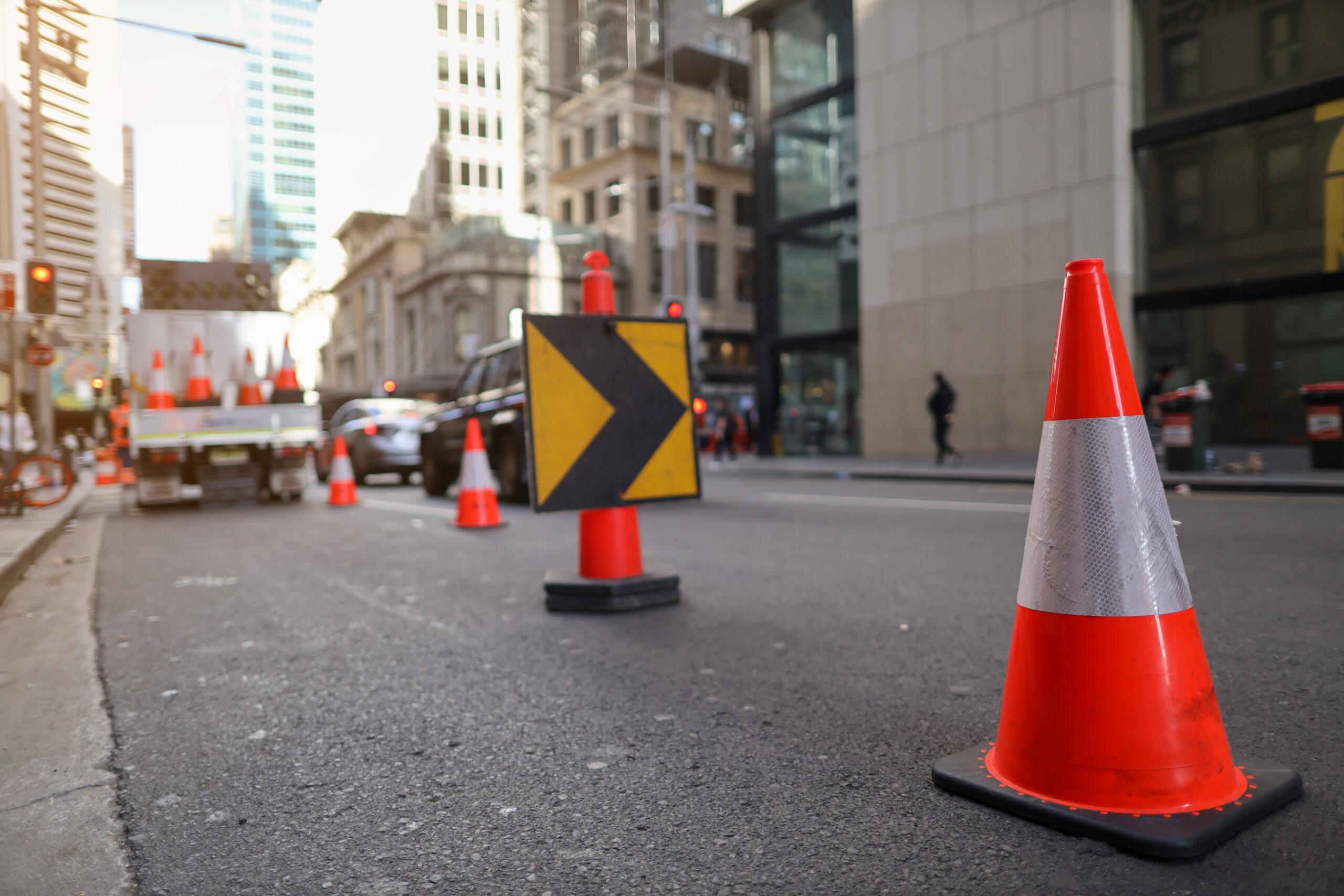 Traffic cones and a directional arrow sign on a city street with construction and high-rise buildings in the background.