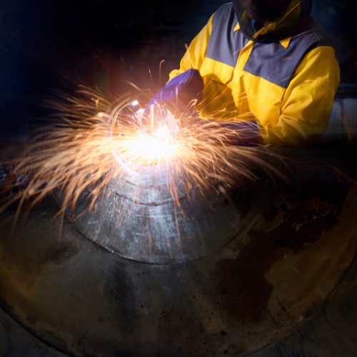 A person wearing protective gear and a yellow jacket is welding metal. Bright sparks fly around the area in a dark environment, highlighting the focus on the welding process.