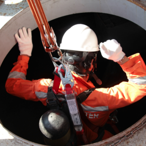 A worker in protective gear, including a hard hat and gloves, descends into a confined space opening using safety harness equipment.