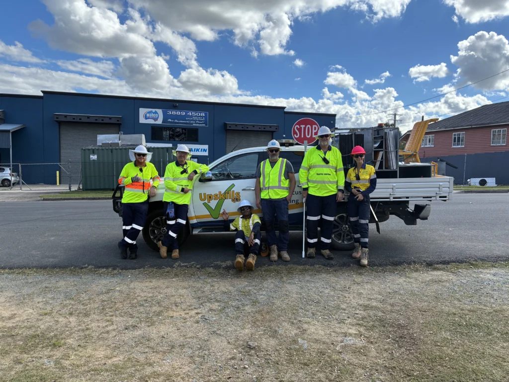 A group of six traffic management trainees and trainers wearing high-visibility workwear and hard hats stand beside an **Upskills QLD** training vehicle during a practical traffic management session. One participant is seated in front of the vehicle while others pose holding traffic control equipment, including a **STOP** sign.  nationally recognised Traffic Control and Traffic Management courses in Queensland.