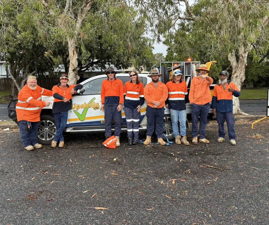 A group of eight traffic management trainees and trainers wearing high-visibility orange work shirts stand beside an **Upskills QLD** training vehicle during a practical TMI (Traffic Management Implementer) training session in **Mackay, Queensland**. The group poses outdoors in a tree-lined training area, with traffic management equipment loaded on the vehicle behind them. The image showcases hands-on, nationally recognised traffic management training in a real-world environment.